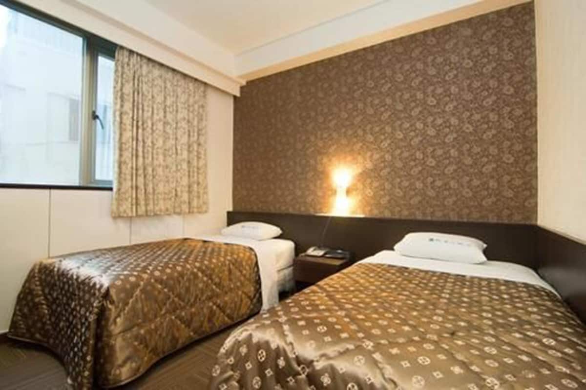 A hotel room with two beds, a nightstand, a lamp, and a window with curtains.