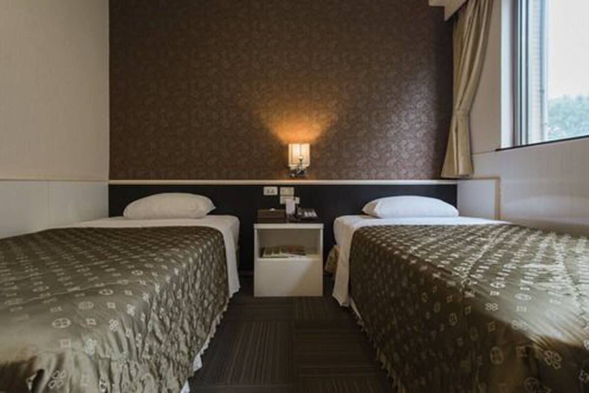 A hotel room with two beds, a nightstand, a wall-mounted lamp, and a window with curtains.