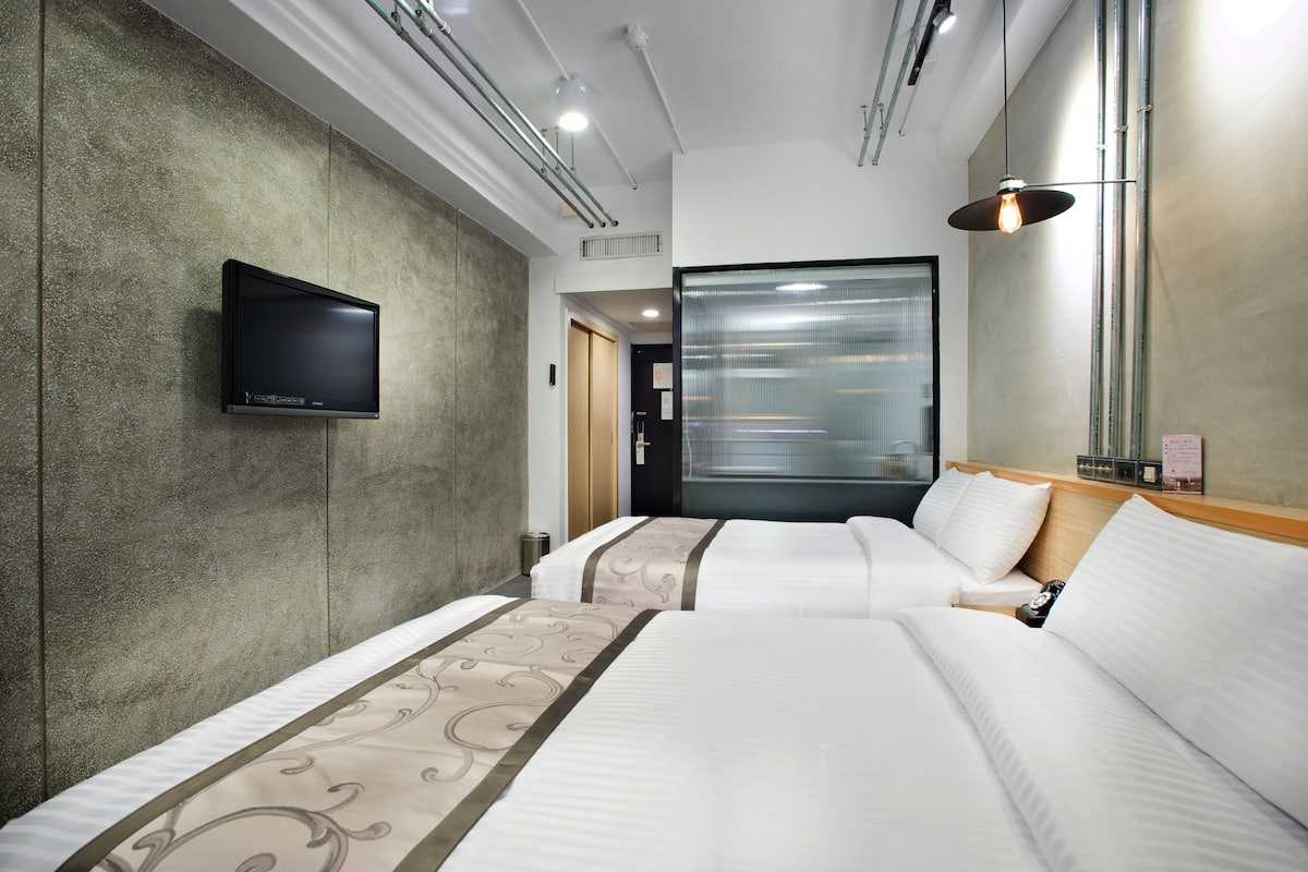 A hotel room with two beds, a flat-screen TV mounted on the wall, and a large window with blinds.