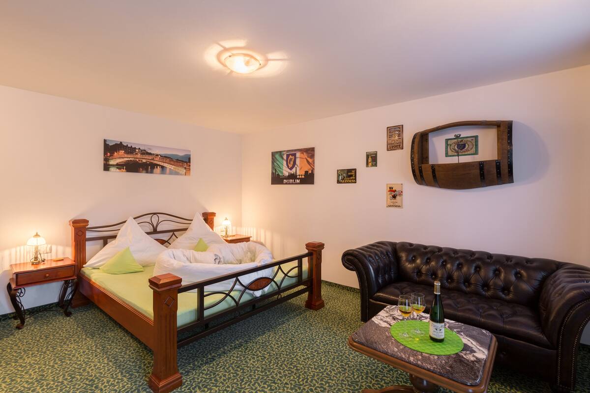 A hotel room with a bed, a leather sofa, a small table with a bottle and glasses, and various wall decorations including a framed picture of a bridge and a barrel-shaped artwork.