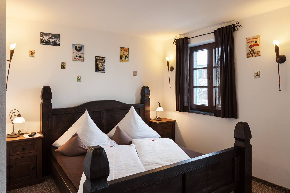 A neatly made bed with white linens and two pillows, a wooden headboard, a nightstand with a lamp, and a window with curtains.