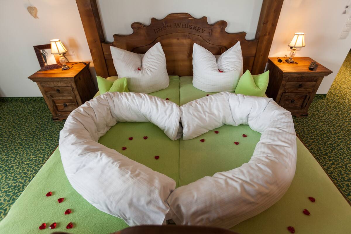 A bed with a heart-shaped pillow arrangement, a wooden headboard with "IRISH WHISKEY" text, and two bedside tables with lamps.