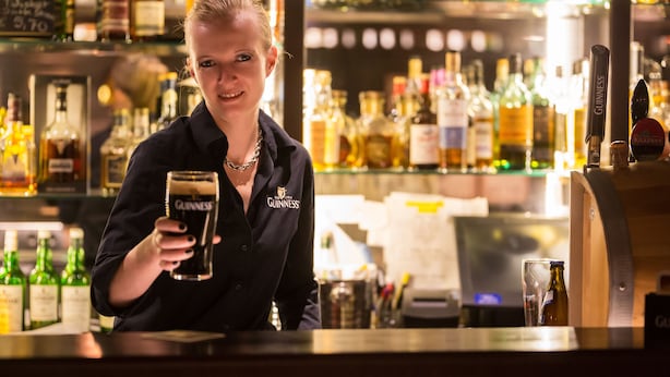 A bartender holding a Guinness beer glass at a bar with a variety of liquor bottles in the background. - Vitusfort (Fuessen)
