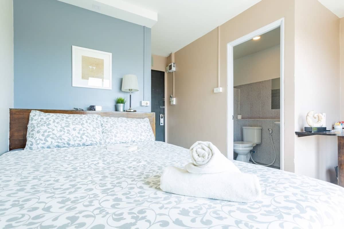 A bedroom with a large bed, a bedside table with a lamp, a framed picture on the wall, and a bathroom visible through an open door.