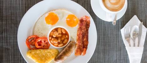 Un plato de desayuno con huevos, tocino, frijoles, salchichas y una rebanada de tostada, acompañado de una taza de café y cubiertos sobre un mantel.