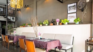 Un restaurant avec un long comptoir, plusieurs tables et des chaises. On peut voir des plantes en pot et un escalier au fond.