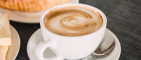 Una taza de café con arte de latte sobre un platito, acompañada por un croissant y una rebanada de pan en un plato.