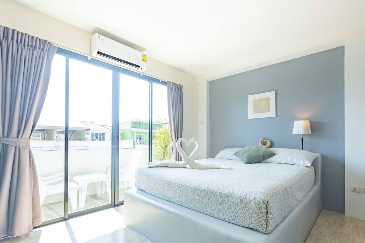 A bedroom with a bed, a window with curtains, a wall-mounted air conditioner, and a bedside lamp.