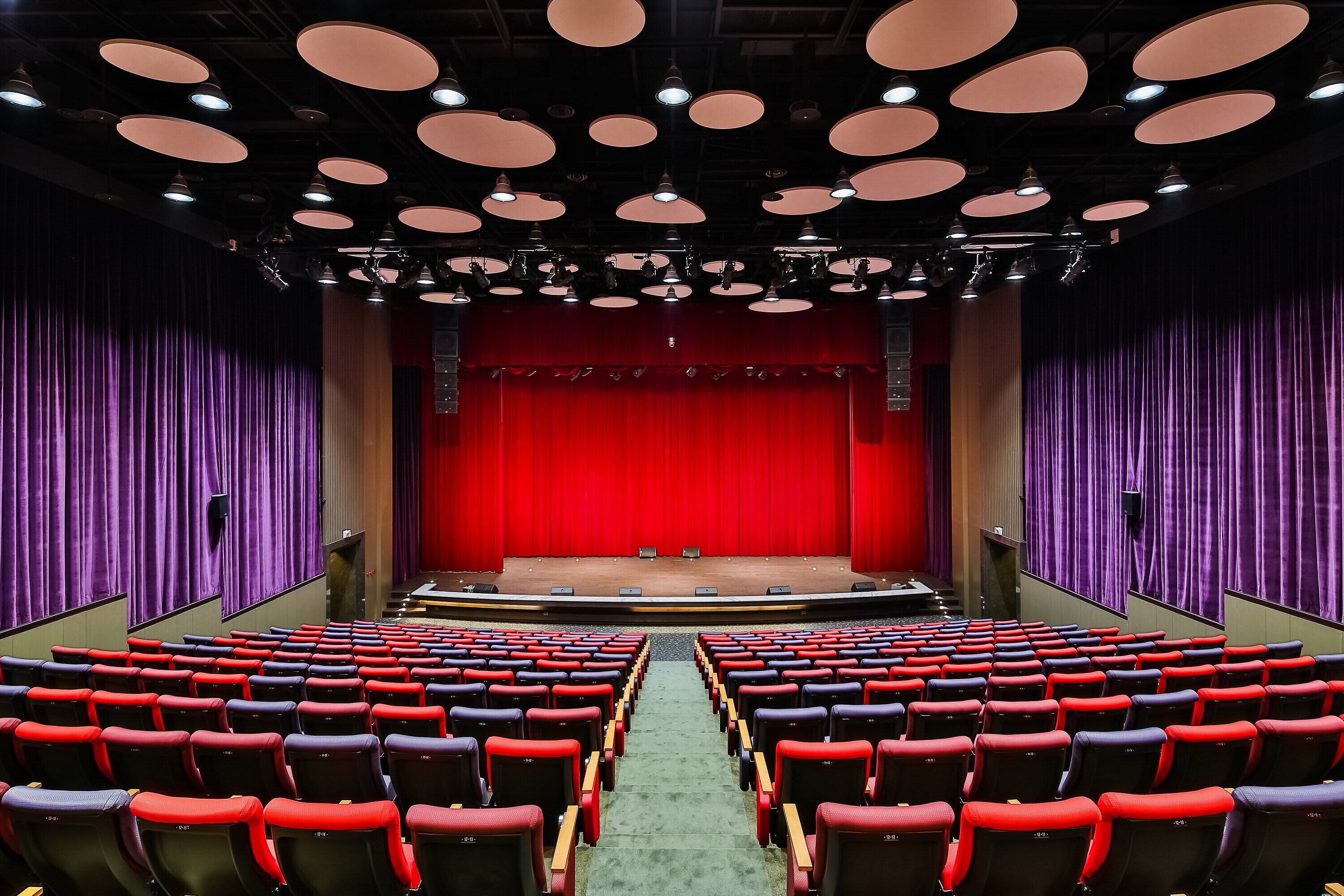 Een auditorium met rijen rode en zwarte stoelen, een centraal podium met een rode gordijn als achtergrond, en meerdere plafondlampen.