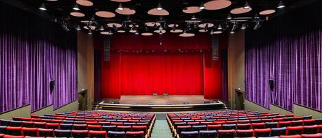 An auditorium with rows of red and black seats, a central stage with a red curtain backdrop, and multiple ceiling lights.