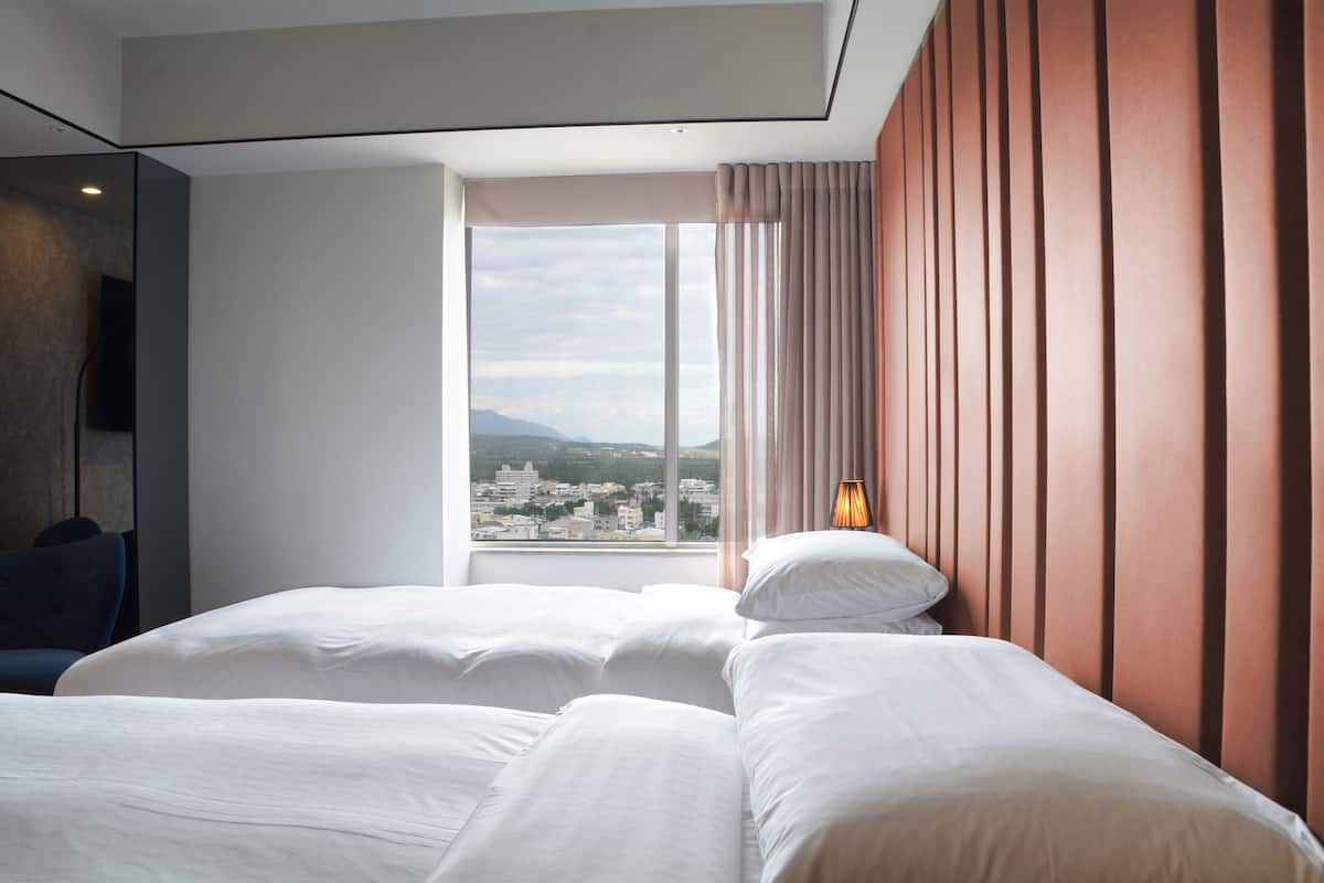 Ein Hotelzimmer mit zwei Betten, einem großen Fenster und Blick auf die Stadt, sowie einem modernen Interieur.