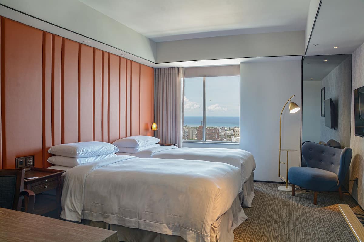 Ein Hotelzimmer mit zwei Betten, einem Schreibtisch, einem Sessel, einer Stehlampe und einem großen Fenster mit Blick auf die Stadt.