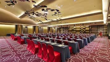 Un salón de conferencias con mesas y sillas dispuestas en filas, una alfombra estampada y un techo decorativo.