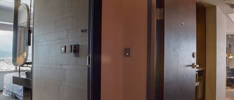 A modern hotel room with a large window, a wooden door, and a view of the cityscape.