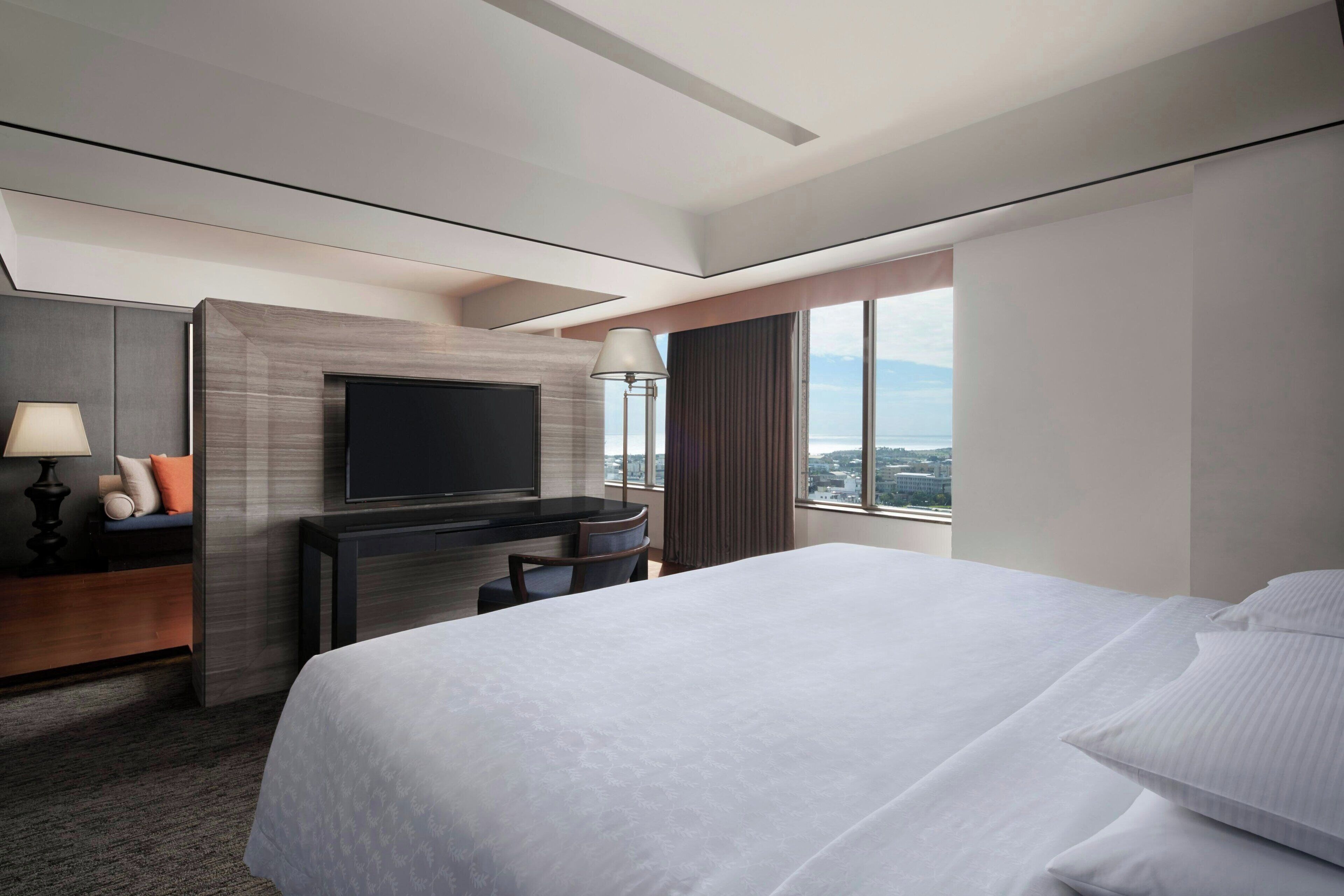 A modern hotel room with a large bed, a flat-screen TV mounted on a wooden panel, a desk with a lamp, and a view of the city through large windows.