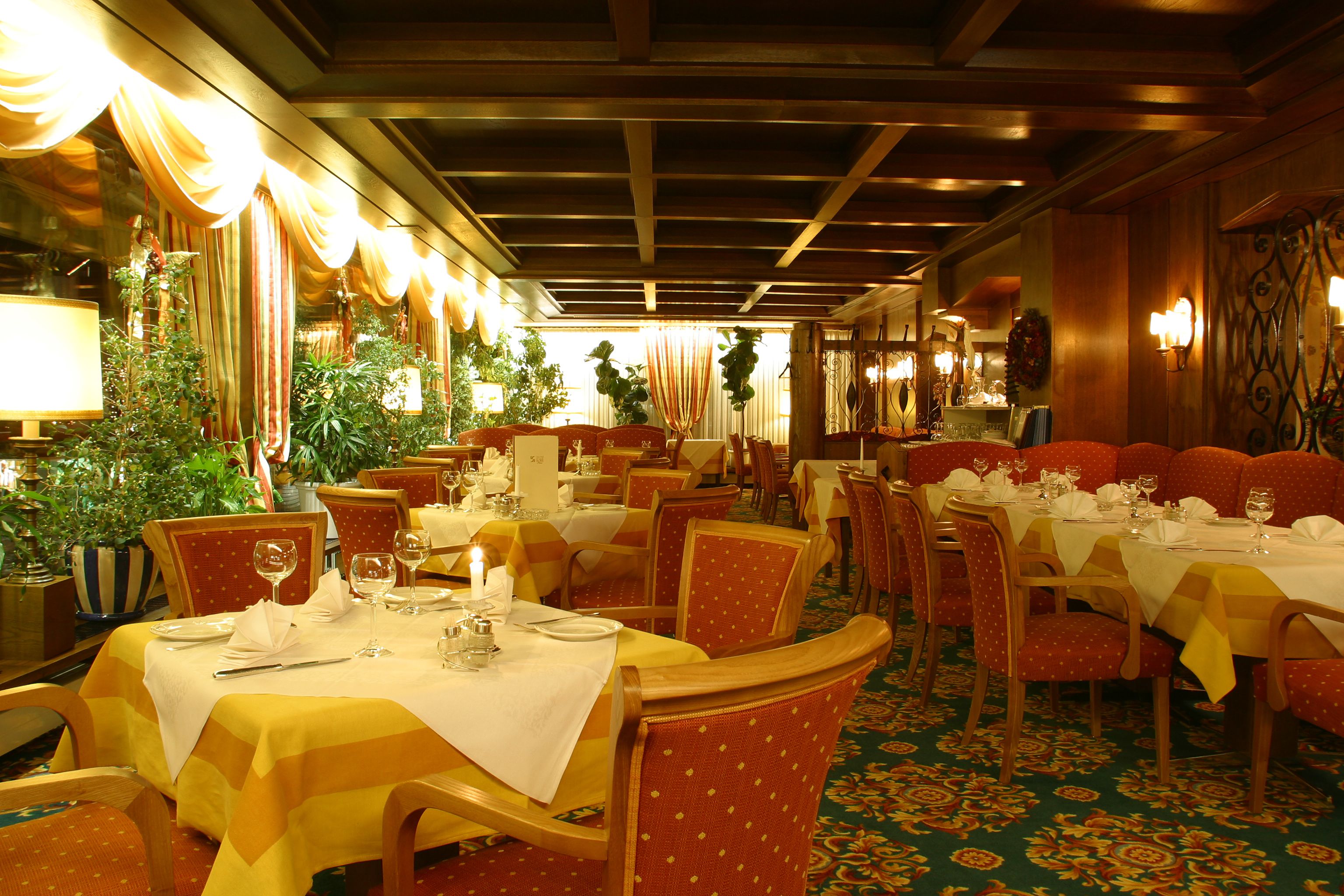 Un restaurant doté de nombreuses tables dressées pour le service, avec un intérieur chaleureux agrémenté de poutres en bois et d’un tapis à motifs.