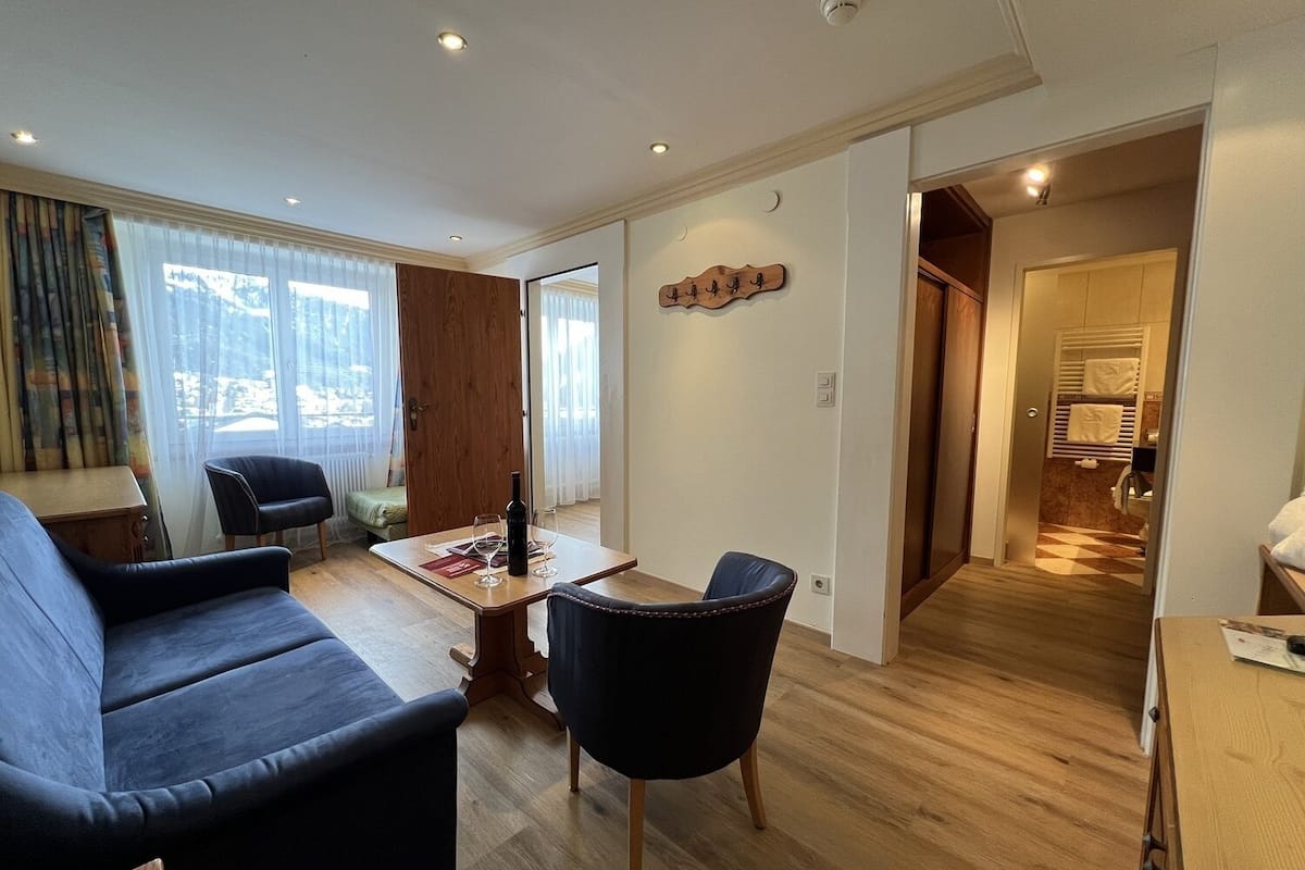 Ein modernes Wohnzimmer mit einer Couch, Sesseln, einem Esstisch und Blick durch ein großes Fenster.