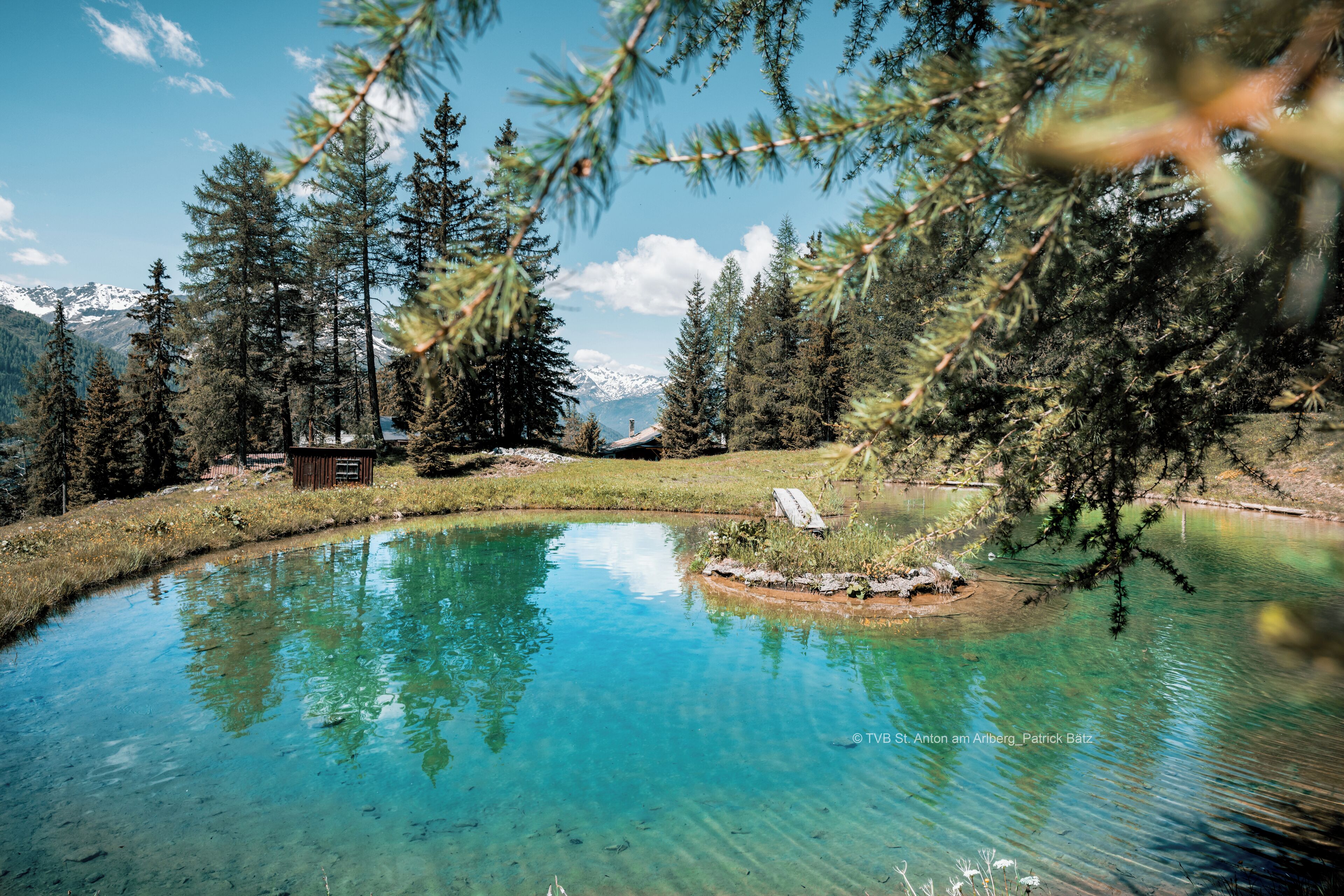 A clear lake surrounded by trees and mountains, with a cabin in the background.