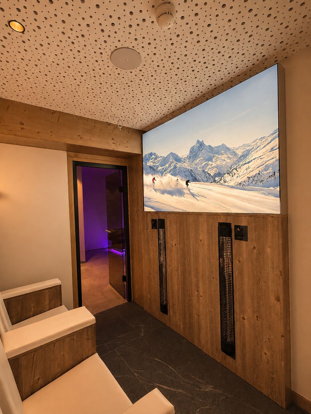 Une pièce dotée d’un grand écran diffusant un paysage de montagne enneigé, de lambris en bois et d’un espace détente avec des fauteuils massants.
