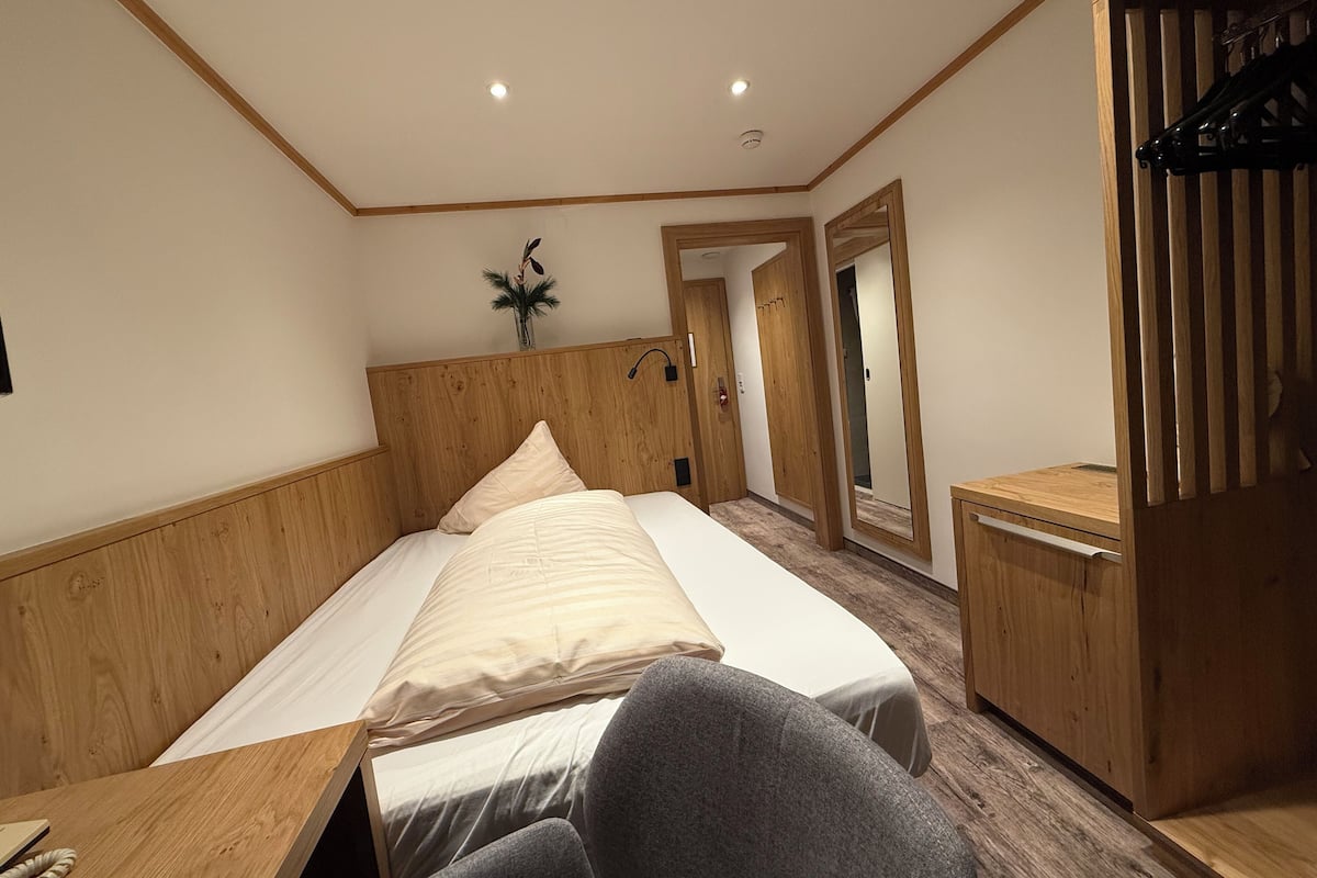 Ein Hotelzimmer mit einem hölzernen Kopfteil, einem Bett mit weißer Bettwäsche, einem kleinen Schreibtisch mit Telefon und einem Spiegel an der Wand.