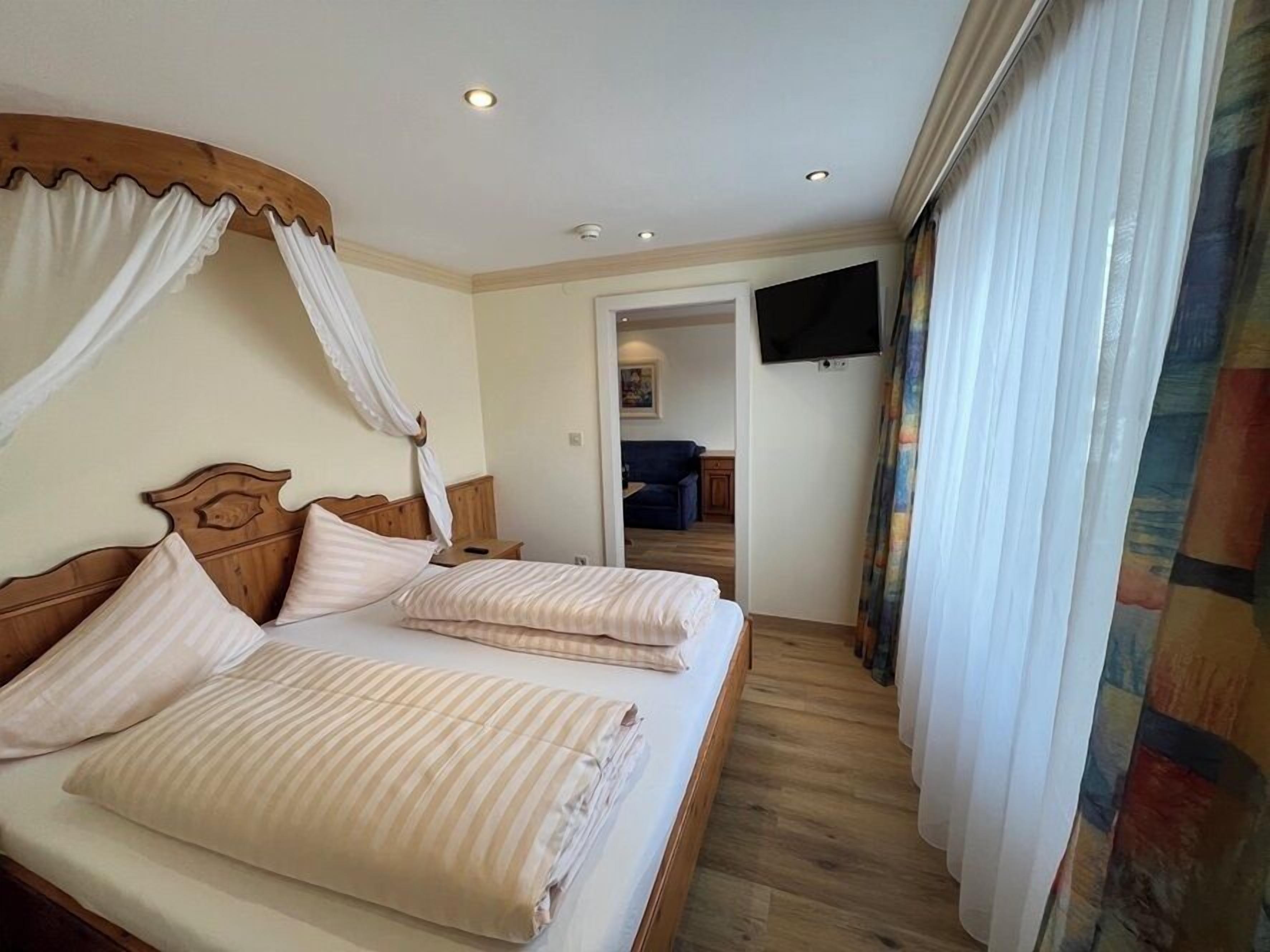 A double bed with a canopy, a television mounted on the wall, and a view of a living area through an open door.