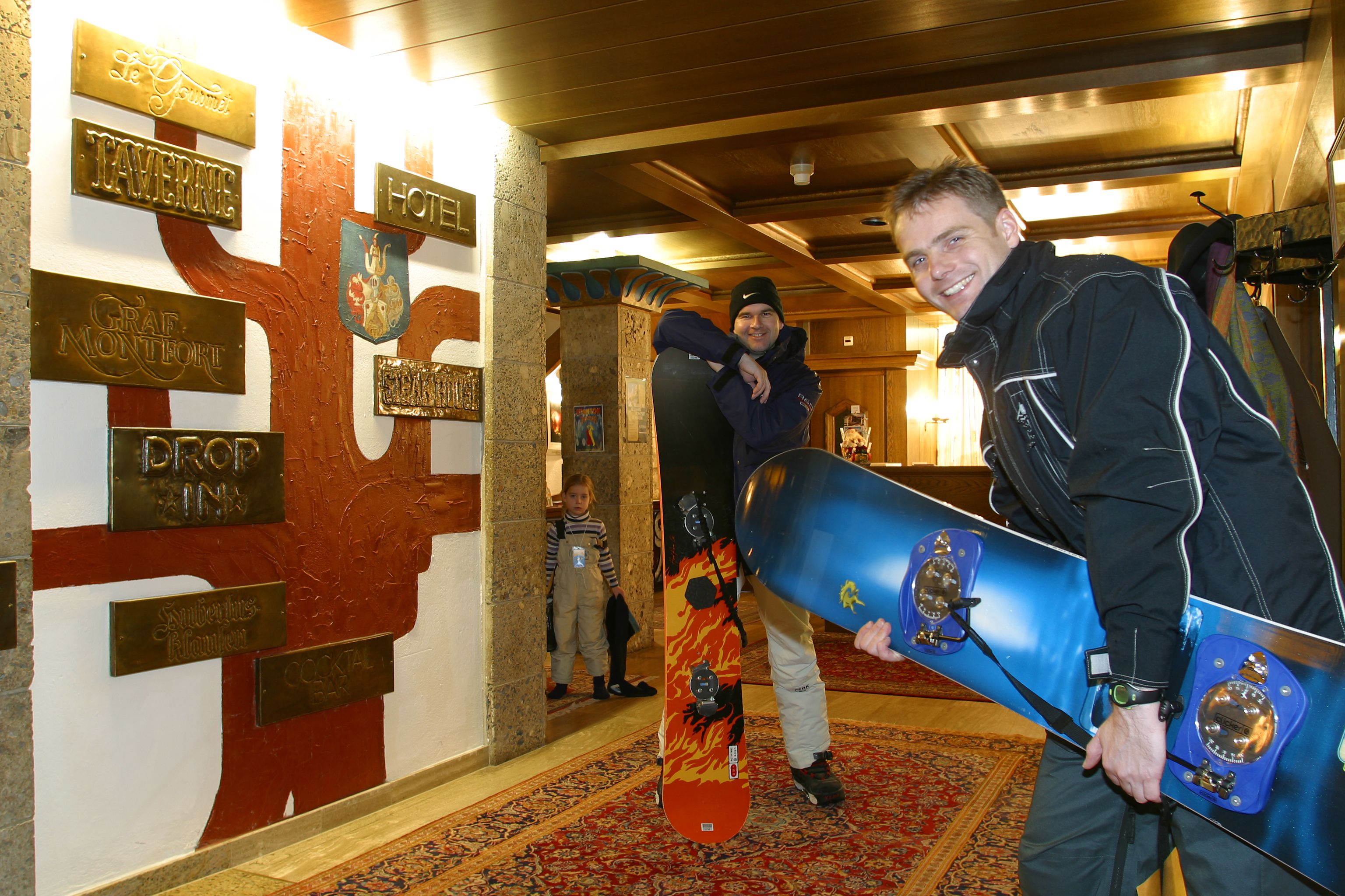 Ein Mann hält ein Snowboard in einer Hotelhalle, wo Schilder verschiedene Annehmlichkeiten und Orte zeigen.