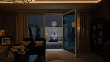 A modern living room with a sofa, armchairs, a coffee table, and a bookshelf. There is a dining area visible through a glass door with a table set for two.