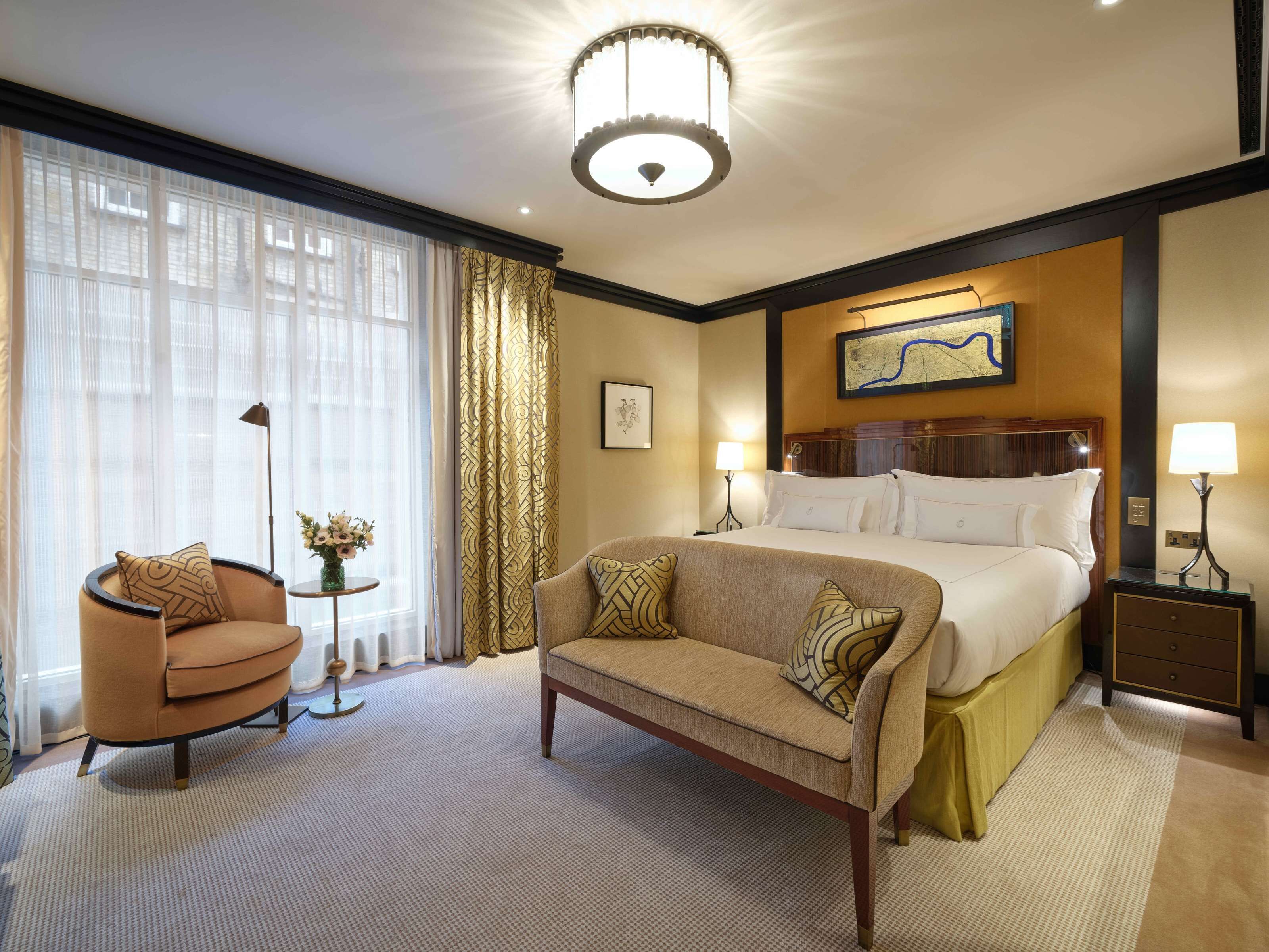 A hotel room with a bed, a sofa, a chair, a nightstand, a lamp, a floor lamp, a map on the wall, and a window with curtains.