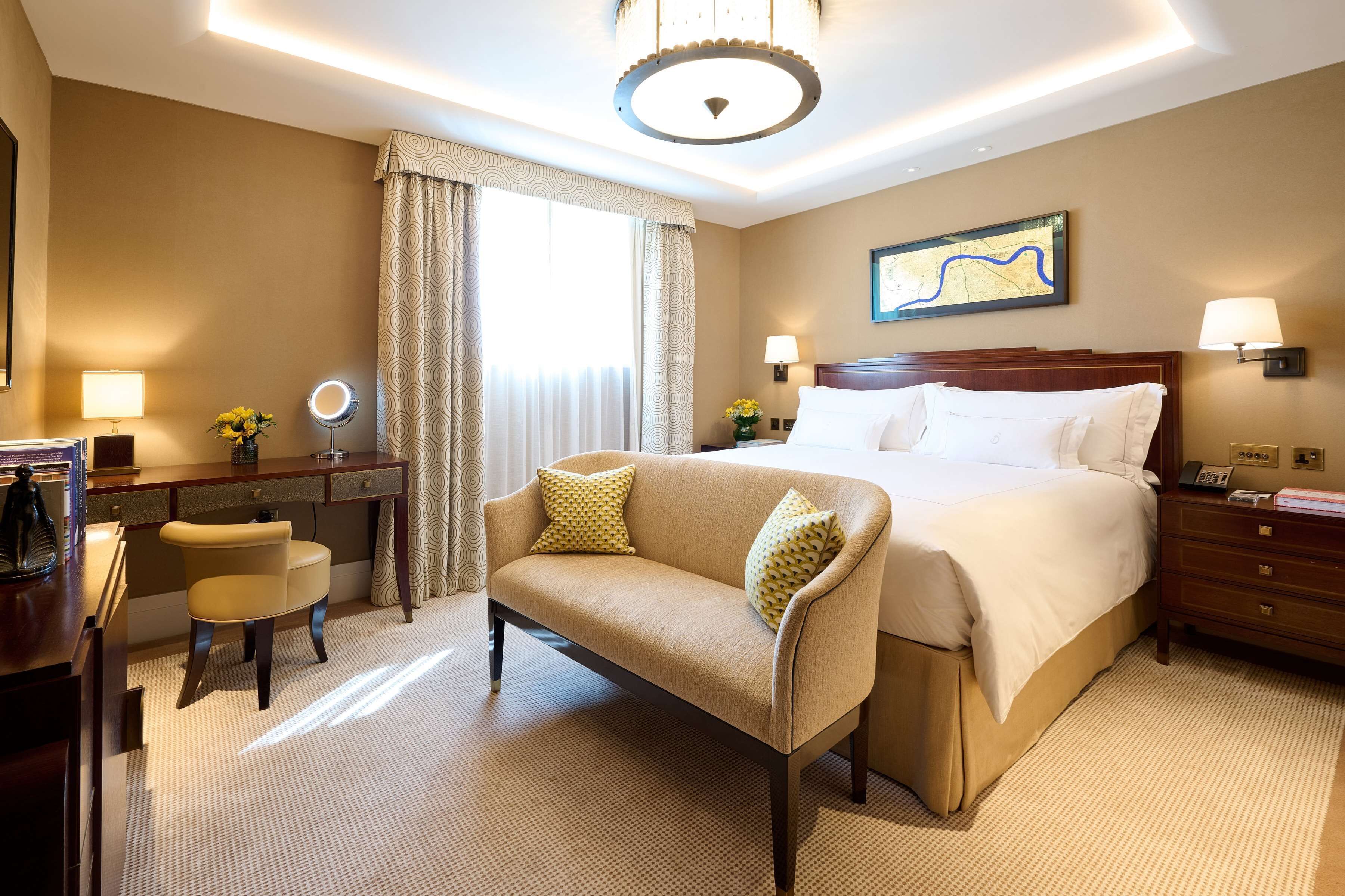 A hotel room with a large bed, a desk with a chair, a small table with a lamp, and a large window with curtains.