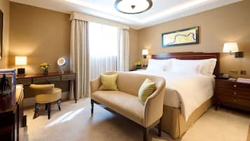 A hotel room with a large bed, a desk with a chair, a small table with a lamp, and a large window with curtains.