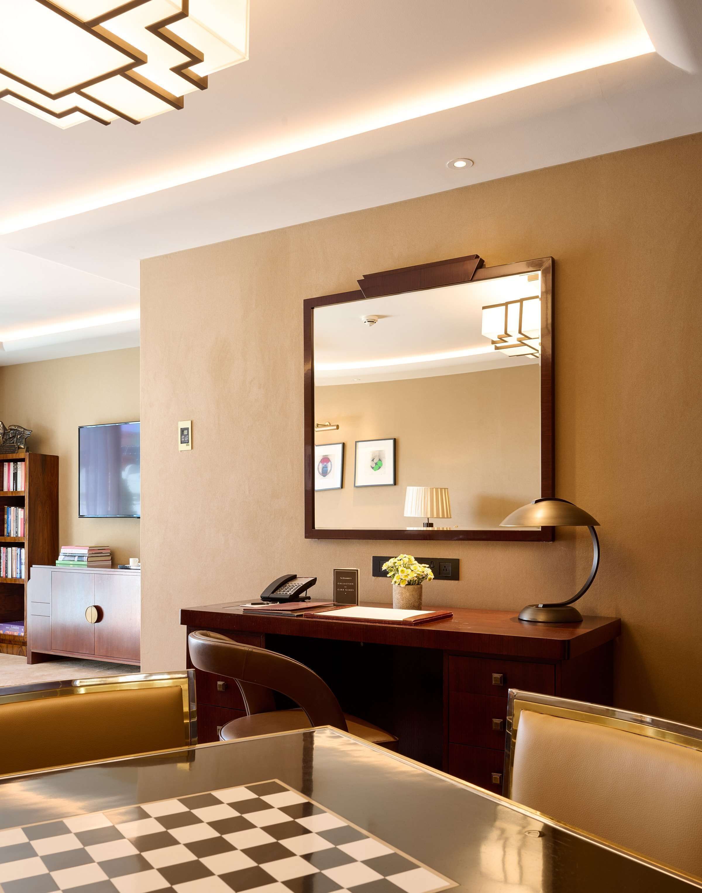 A hotel room with a desk, chair, and a large mirror. There is a bookshelf, a television, and a lamp on the desk.
