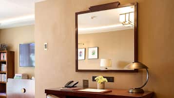 Une chambre d’hôtel avec un bureau, une chaise et un grand miroir. On y trouve également une bibliothèque, un téléviseur et une lampe sur le bureau.