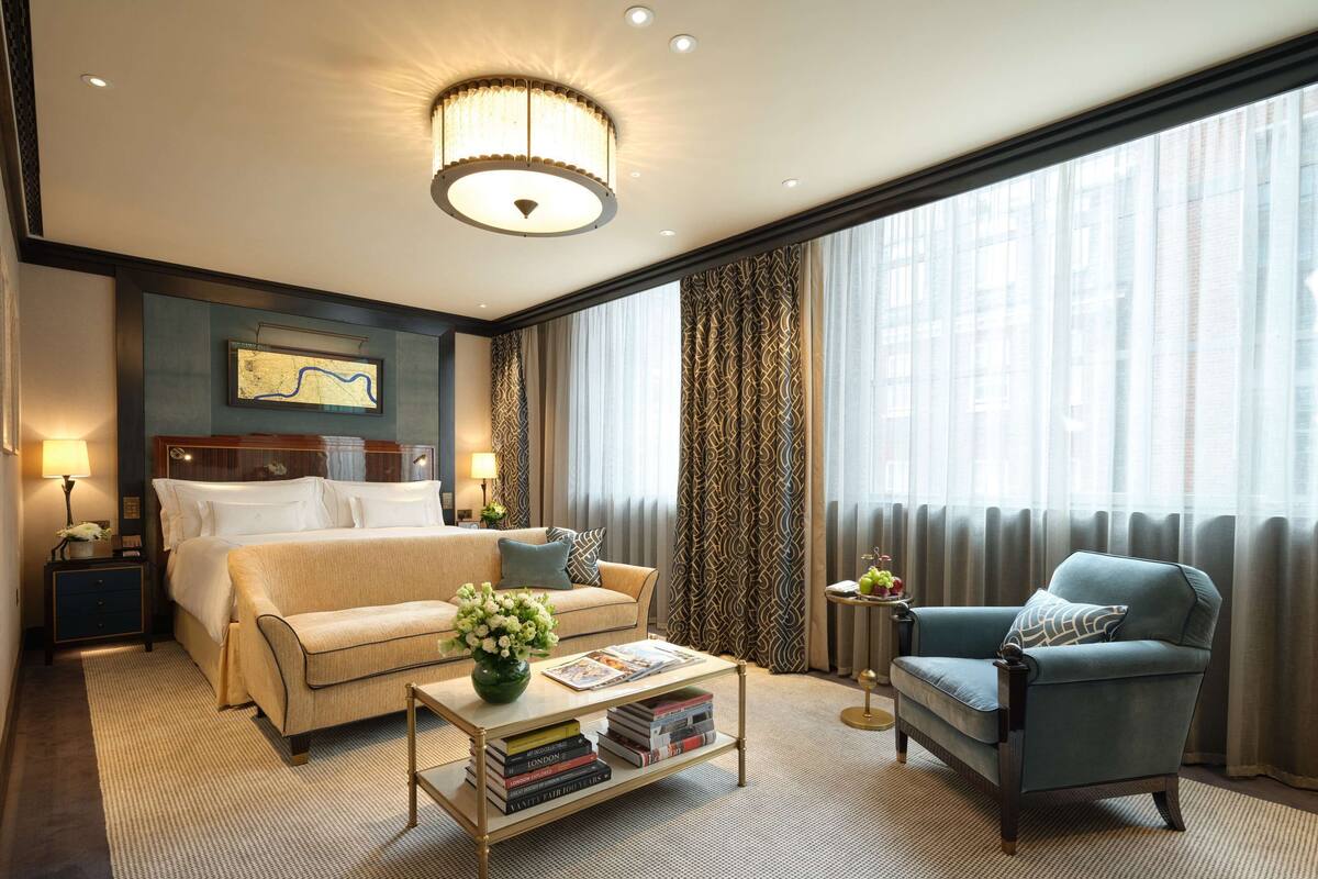 Ein Hotelzimmer mit einem Bett, einem Sofa, einem Sessel, einem Couchtisch mit Büchern und einem großen Fenster mit Vorhängen.