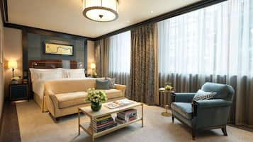 Habitación de hotel con cama, sofá, sillón, mesita de centro con libros y una ventana grande con cortinas.
