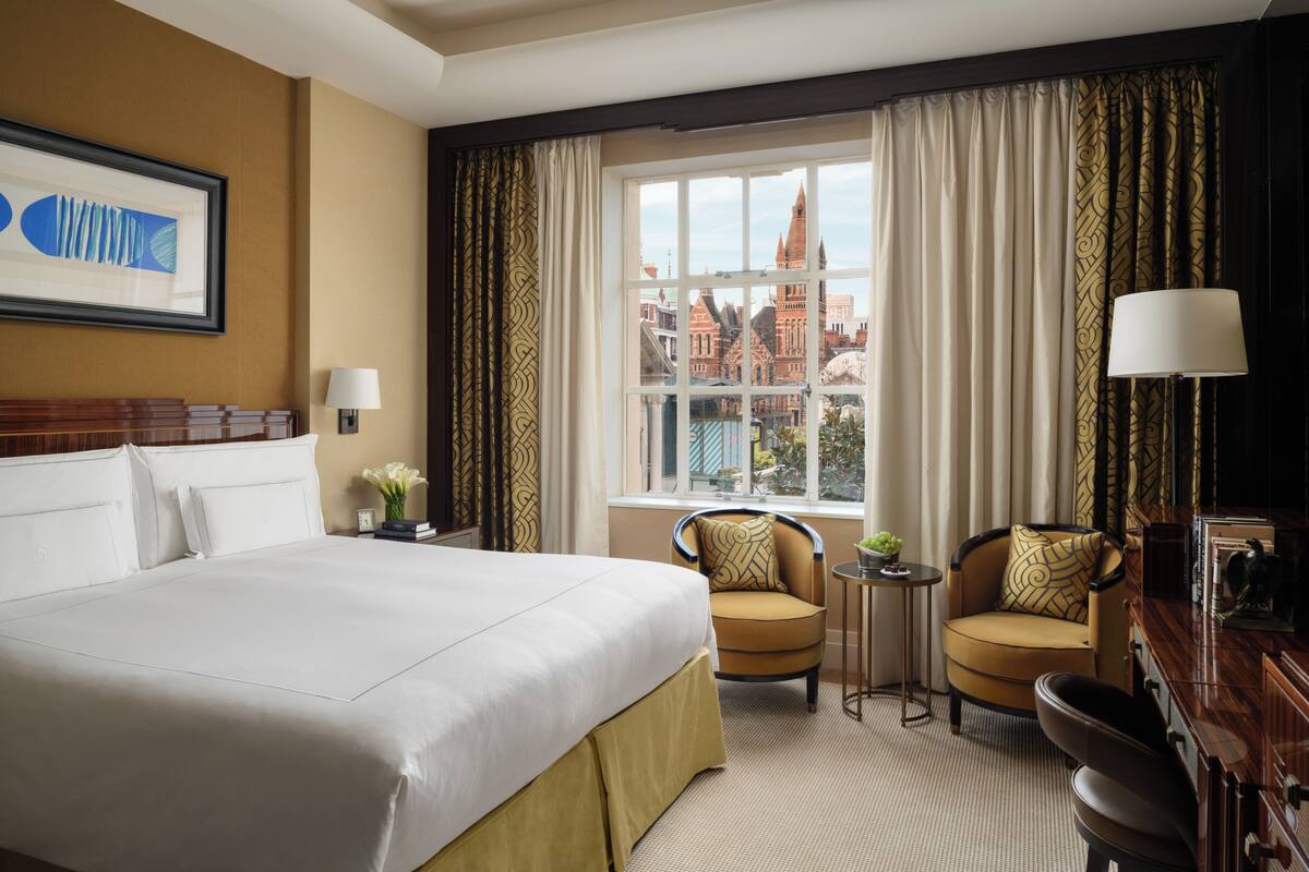 Ein Hotelzimmer mit einem großen Bett, zwei Sesseln, einem kleinen Tisch und Blick auf eine Stadtlandschaft durch ein mit Vorhängen drapiertes Fenster.
