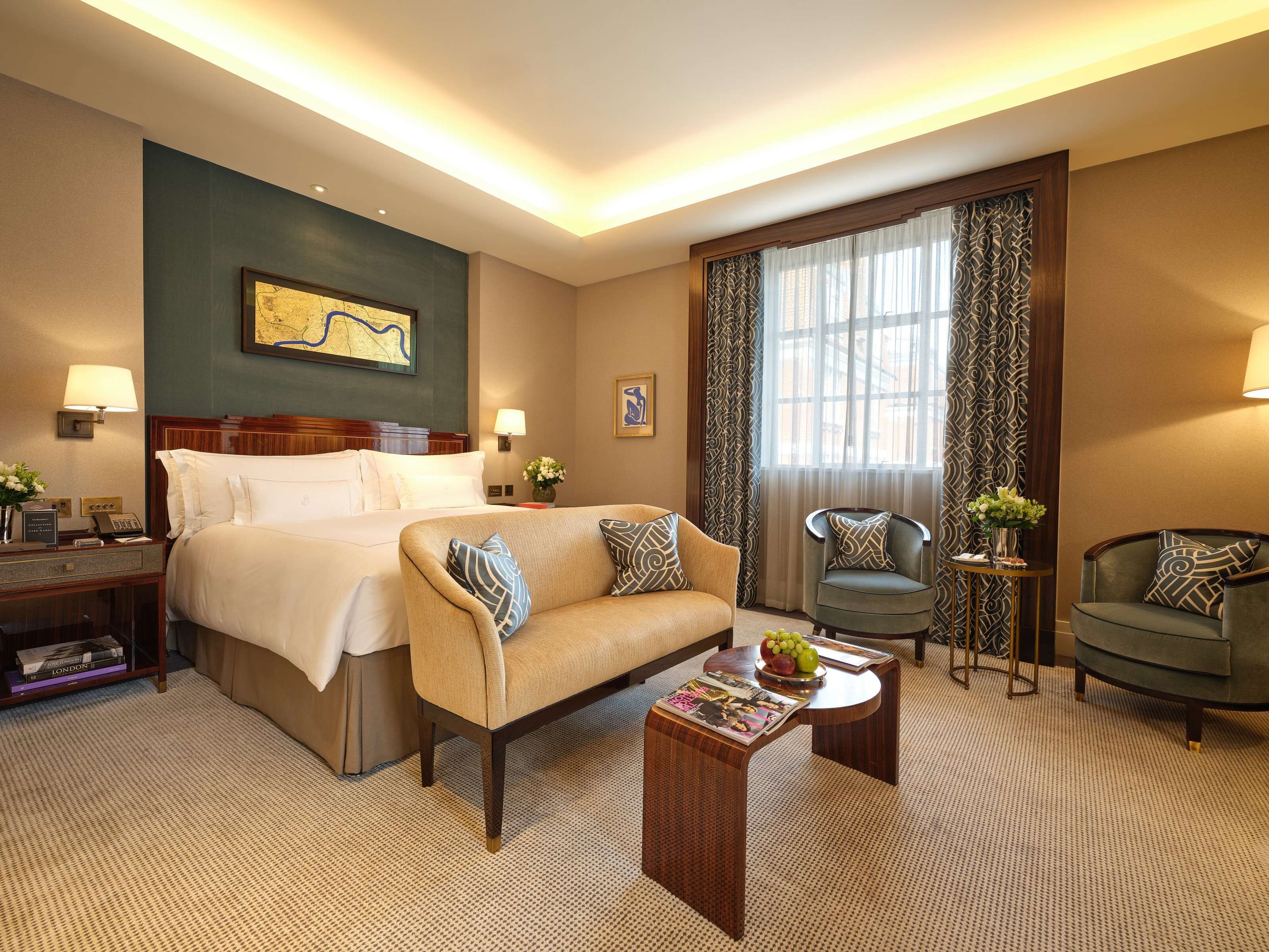 Une chambre d’hôtel avec un grand lit, un canapé, deux fauteuils, une petite table sur laquelle se trouve un panier de fruits, et une grande fenêtre avec des rideaux.