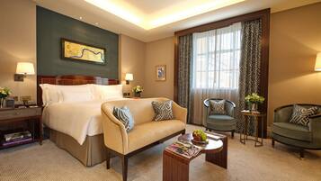A hotel room with a large bed, a sofa, two armchairs, a small table with a fruit bowl, and a large window with curtains.