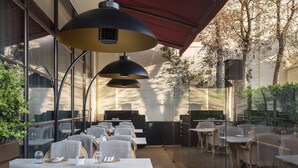An outdoor dining area with wicker chairs and tables, large pendant lights, and a glass enclosure. - The Beaumont Mayfair (London)
