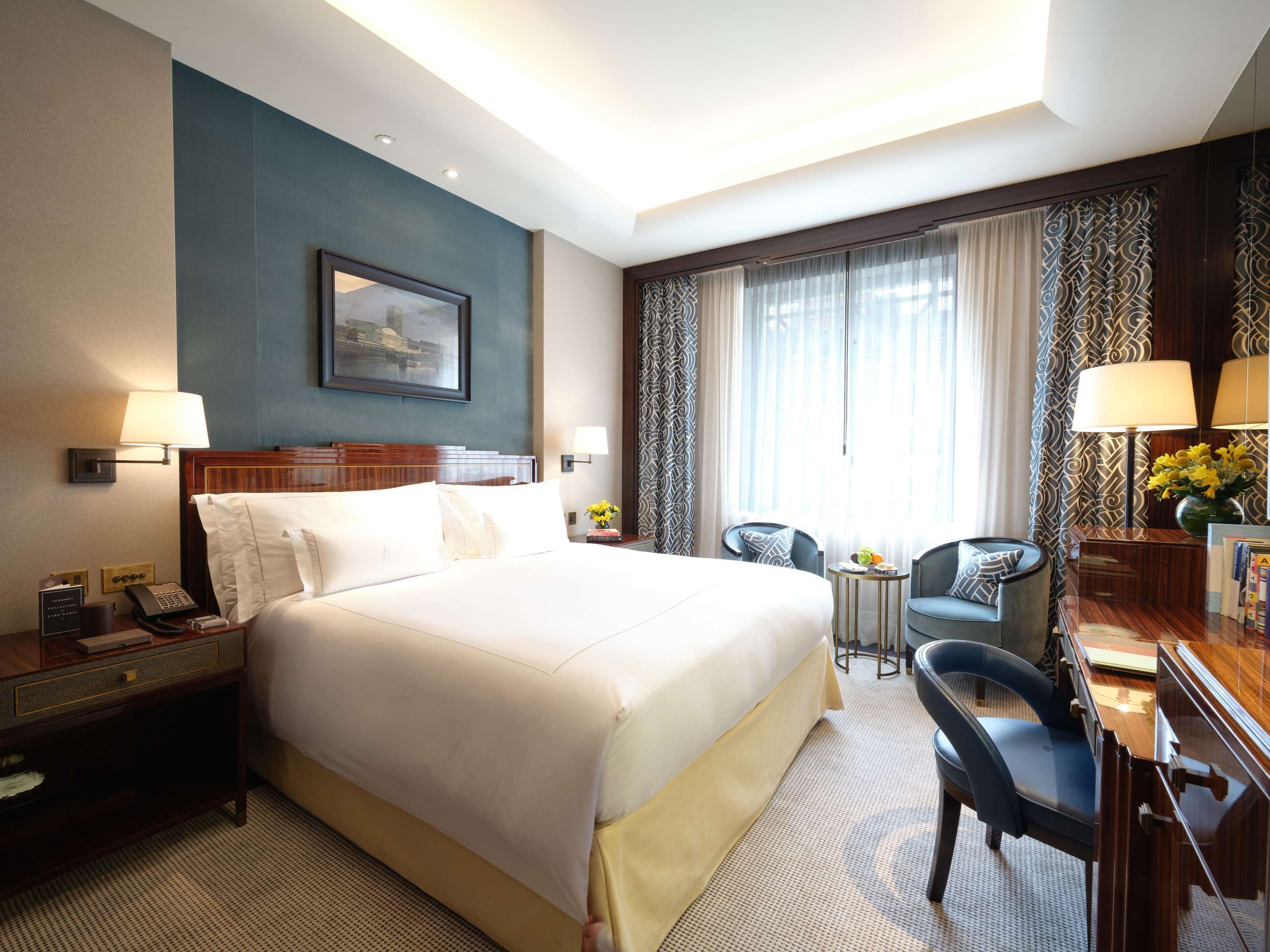 A hotel room with a large bed, bedside tables, a desk with a chair, a small table with a chair, and a window with curtains.