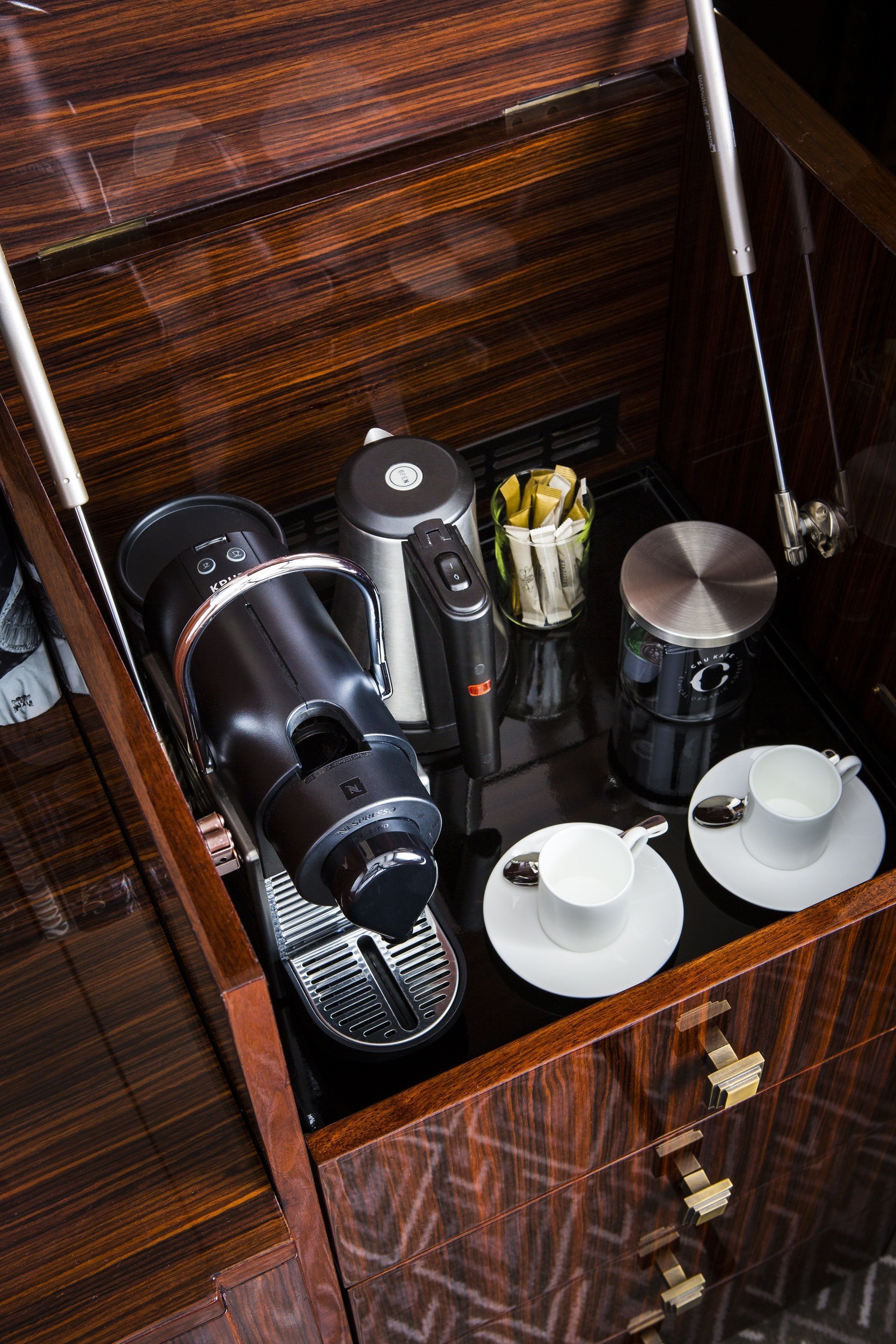 A compact coffee station with a coffee maker, a kettle, and a cup warmer, along with two white cups and saucers.