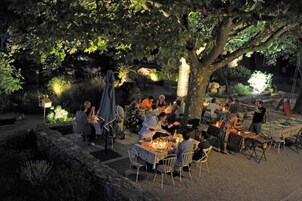 Un espacio al aire libre para comer con mesas y sillas bajo un árbol grande, rodeado de muros de piedra y follaje.