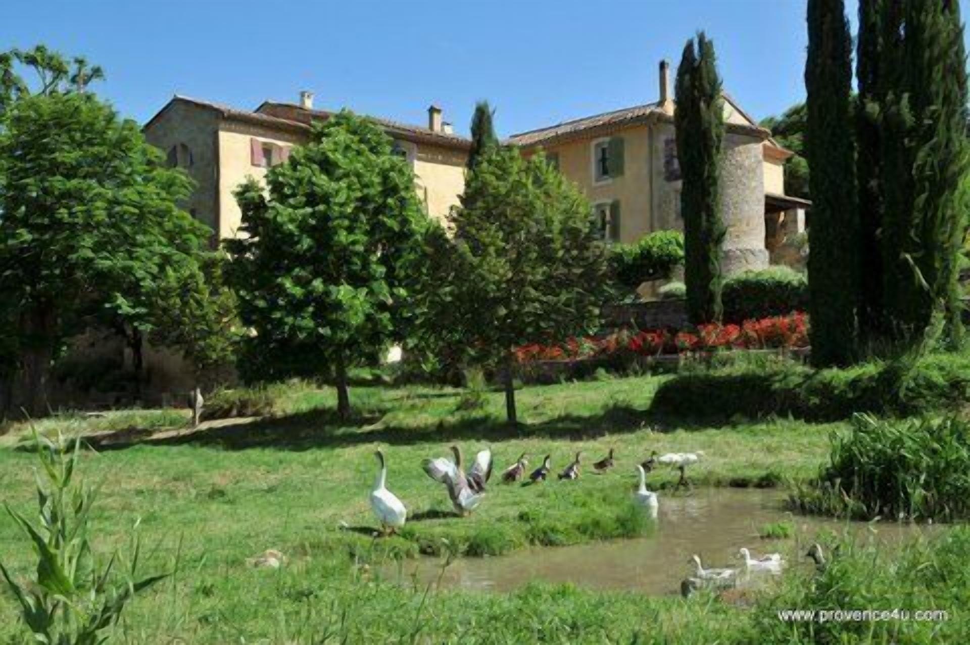 Um jardim tranquilo com um lago, rodeado por árvores e um edifício com telhado em azulejos.