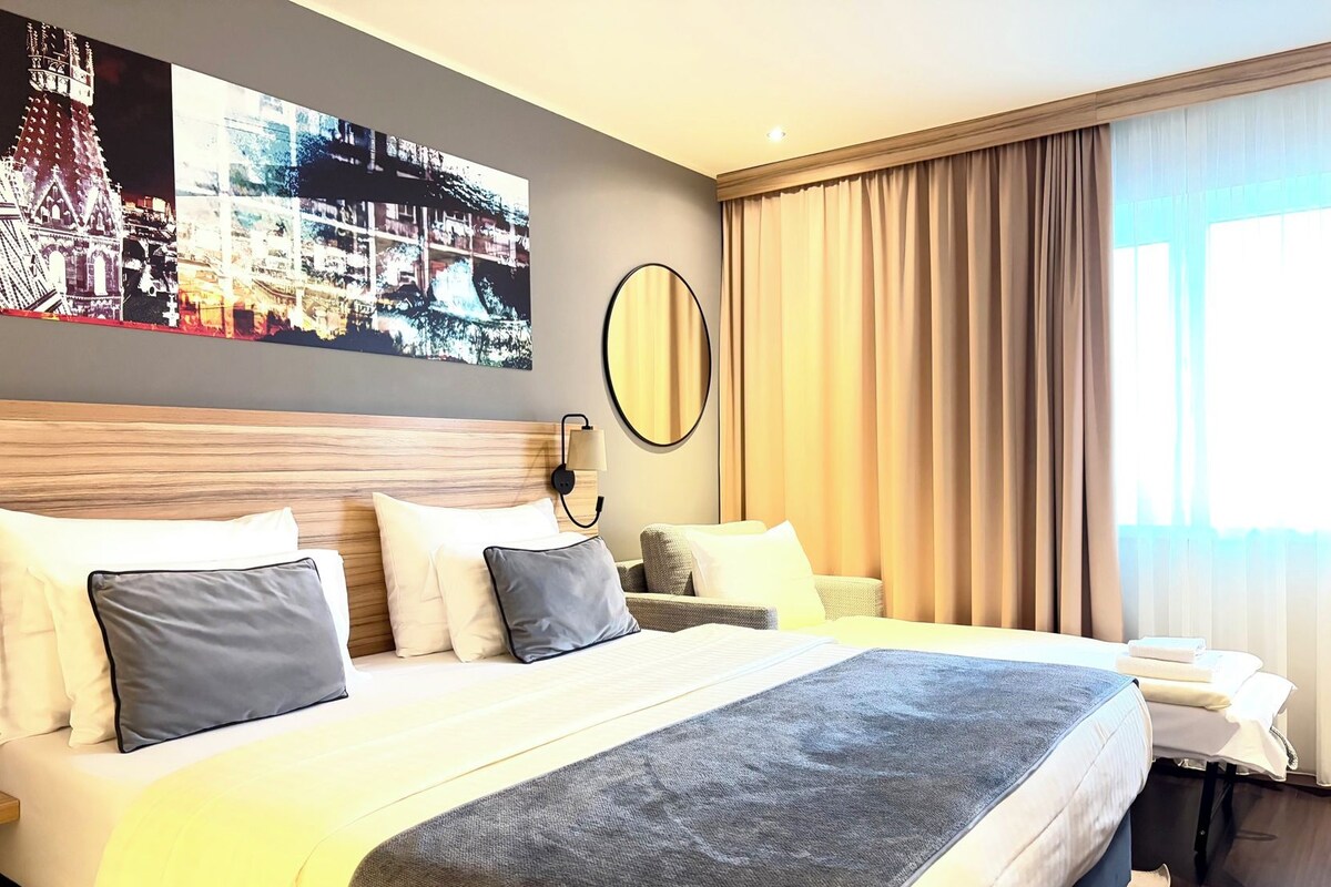 A hotel room with a large bed, a wall-mounted mirror, and a window with curtains.