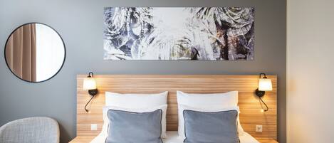Una habitación de hotel con una cama, dos lámparas de noche, un espejo redondo, una silla y un cuadro abstracto de gran tamaño en la pared.