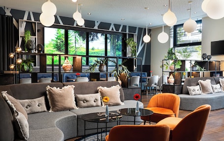 A modern lounge area with sofas, chairs, and a coffee table. Large windows provide a view of greenery outside.