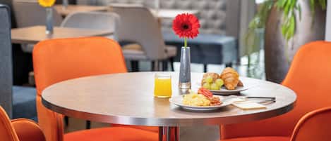 A dining area with orange chairs and a round table, featuring breakfast items like eggs, toast, and a croissant.