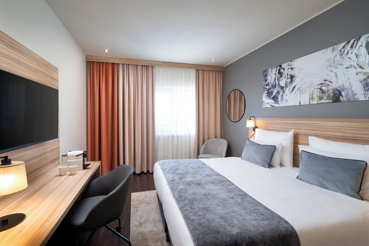 Ein modernes Hotelzimmer mit einem großen Bett, einem Schreibtisch mit Lampe, einem Sessel, einem Flachbildfernseher und einem Bild an der Wand.