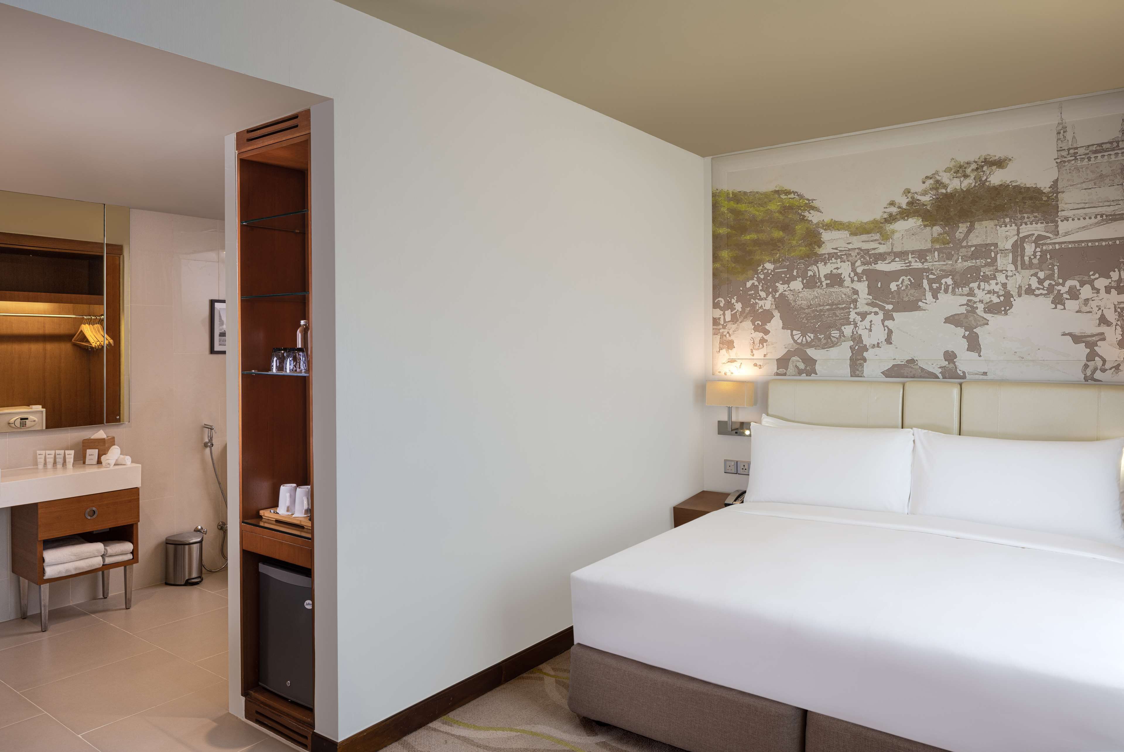 A hotel room with a bed, a wooden wardrobe, a bathroom with a mirror, and a wall with a historical mural.