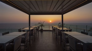 Un área de comedor al aire libre con mesas y sillas, con vista al mar al atardecer.