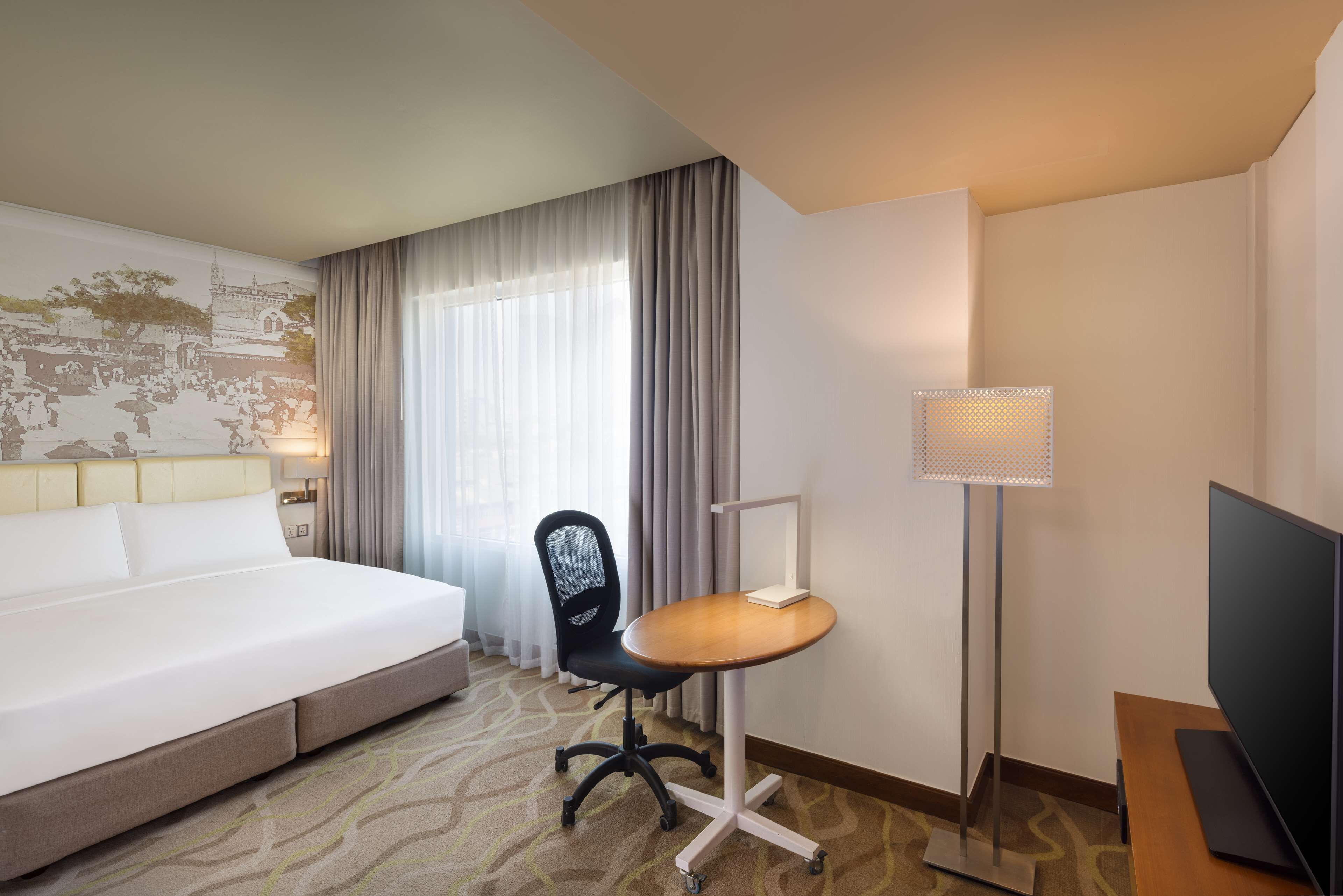 A hotel room with a large bed, a desk with a chair, a TV, a lamp, and a world map on the wall.